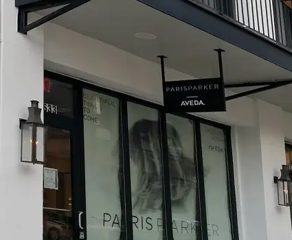 - Paris Parker Aveda Salons and Spas - Baton Rouge, New Orleans, Mandeville, LA Storefront of Paris Parker Aveda Salon with glass windows, exterior lights, and a hanging black sign above the entrance—your destination for a relaxing haircut or rejuvenating facial. - Paris Parker Aveda Salons and Spas - Baton Rouge, New Orleans, Mandeville, LA