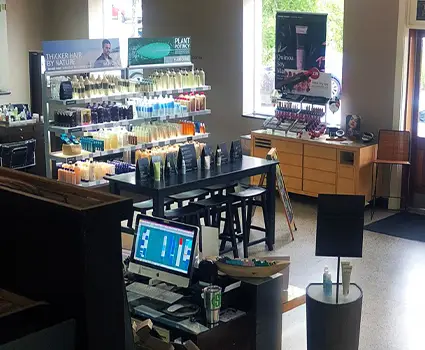 - Paris Parker Aveda Salons and Spas - Baton Rouge, New Orleans, Mandeville, LA Interior of an Aveda Salon with shelves stocked with hair products, a table displaying items, a computer at the front desk, and natural light streaming in—perfect for your next balayage appointment. - Paris Parker Aveda Salons and Spas - Baton Rouge, New Orleans, Mandeville, LA