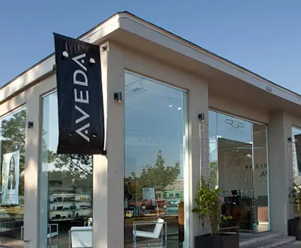 - Paris Parker Aveda Salons and Spas - Baton Rouge, New Orleans, Mandeville, LA Exterior view of a modern Aveda Salon with large glass windows, displaying an "AVEDA" banner and minimalistic outdoor seating—offering balayage and day spa services. - Paris Parker Aveda Salons and Spas - Baton Rouge, New Orleans, Mandeville, LA