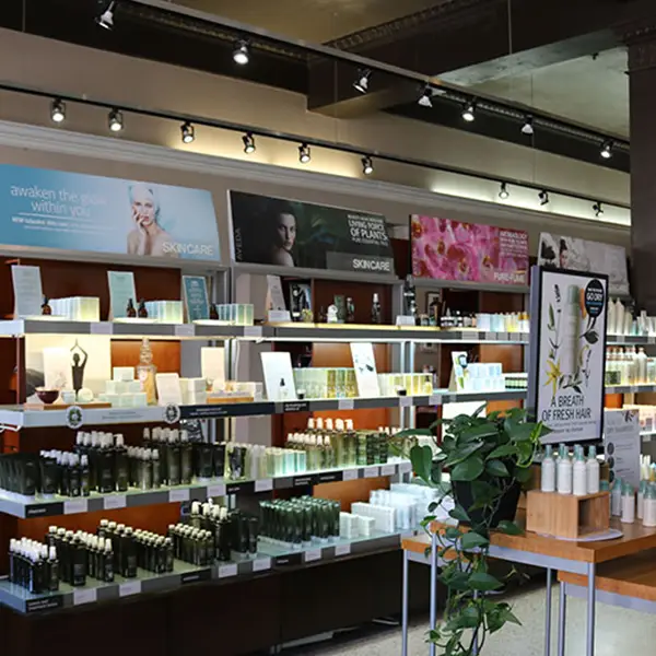 - Paris Parker Aveda Salons and Spas - Baton Rouge, New Orleans, Mandeville, LA Interior of a skincare store with shelves displaying various bottles and products, promotional posters on the wall, and a small plant on a wooden table in the foreground—similar to a serene Aveda Salon or facial spa. - Paris Parker Aveda Salons and Spas - Baton Rouge, New Orleans, Mandeville, LA