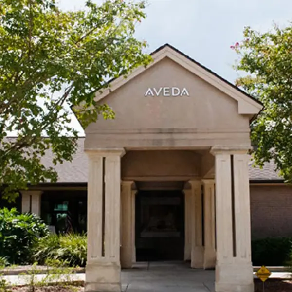 - Paris Parker Aveda Salons and Spas - Baton Rouge, New Orleans, Mandeville, LA Entrance to a building with a peaked roof labeled "AVEDA," surrounded by trees and greenery, inviting guests in for a luxurious facial or expert haircut. - Paris Parker Aveda Salons and Spas - Baton Rouge, New Orleans, Mandeville, LA