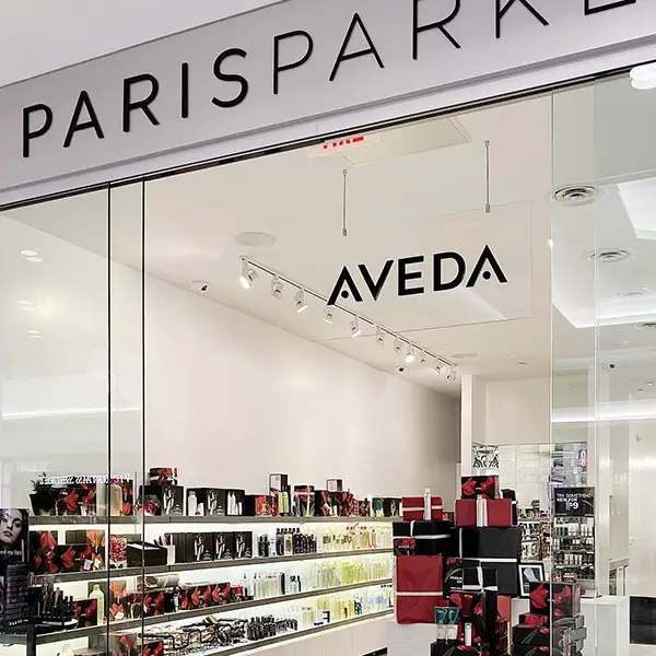 - Paris Parker Aveda Salons and Spas - Baton Rouge, New Orleans, Mandeville, LA A view of a PARISPARKE store entrance with an AVEDA sign inside, displaying shelves stocked with beauty and hair care products for the ultimate Day Spa or Hair Salon experience. - Paris Parker Aveda Salons and Spas - Baton Rouge, New Orleans, Mandeville, LA