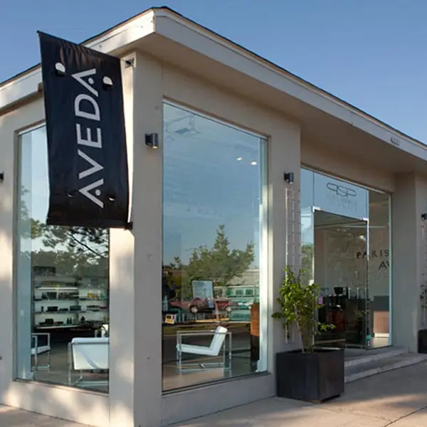 - Paris Parker Aveda Salons and Spas - Baton Rouge, New Orleans, Mandeville, LA Exterior view of a modern Aveda Salon and Day Spa with large glass windows and an AVEDA banner hanging near the entrance. - Paris Parker Aveda Salons and Spas - Baton Rouge, New Orleans, Mandeville, LA