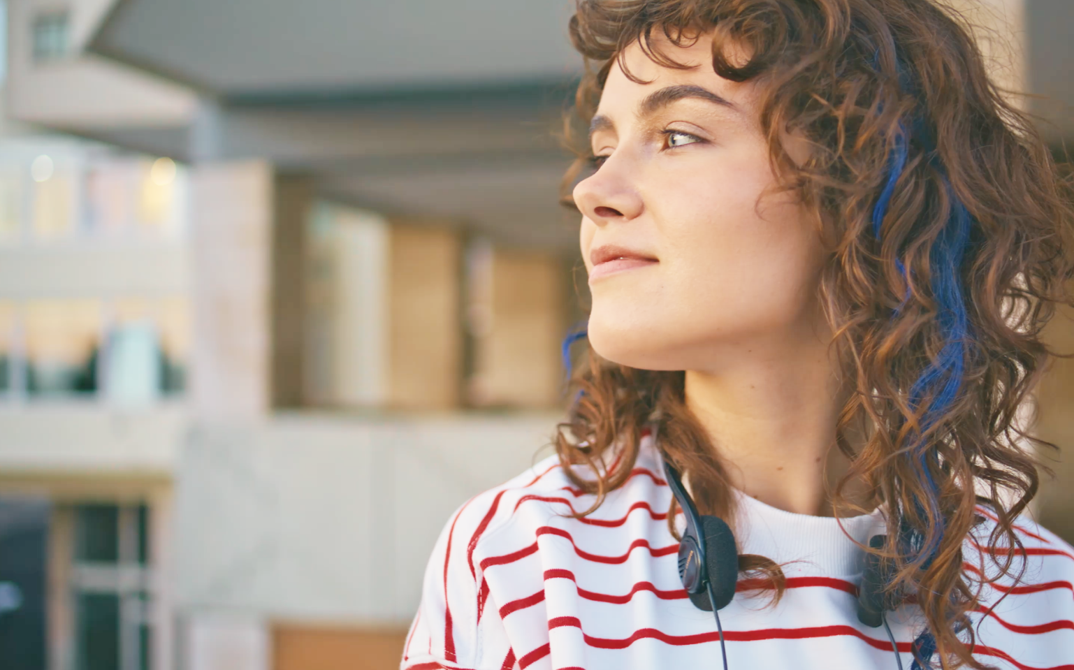 - Paris Parker Aveda Salons and Spas - Baton Rouge, New Orleans, Mandeville, LA A young person with curly hair and headphones around their neck looks thoughtfully to the side, wearing a red and white striped shirt in an outdoor urban setting, perhaps just leaving an Aveda Salon. - Paris Parker Aveda Salons and Spas - Baton Rouge, New Orleans, Mandeville, LA