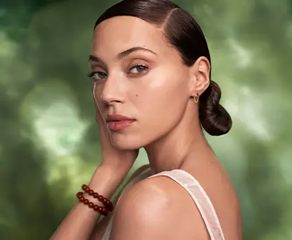 - Paris Parker Aveda Salons and Spas - Baton Rouge, New Orleans, Mandeville, LA Woman with slicked-back dark hair in a low bun, wearing a white top and red beaded bracelet, poses against a green blurred background, touching her face lightly—evoking the serene glow of an Aveda Salon facial. - Paris Parker Aveda Salons and Spas - Baton Rouge, New Orleans, Mandeville, LA