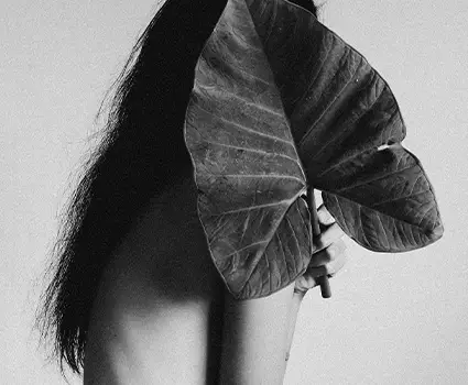 - Paris Parker Aveda Salons and Spas - Baton Rouge, New Orleans, Mandeville, LA A person with long hair, fresh from an Aveda Salon haircut, partially covers their face and upper body with a large leaf, set against a plain background in black and white. - Paris Parker Aveda Salons and Spas - Baton Rouge, New Orleans, Mandeville, LA