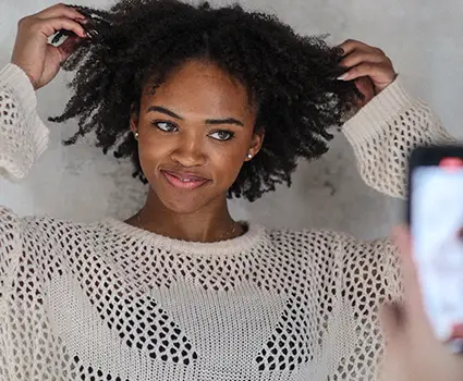 - Paris Parker Aveda Salons and Spas - Baton Rouge, New Orleans, Mandeville, LA Woman in a cream-colored, open-knit sweater adjusts her curly haircut while someone takes a photo of her with a smartphone, capturing the fresh style she got at the Aveda Salon and Day Spa. - Paris Parker Aveda Salons and Spas - Baton Rouge, New Orleans, Mandeville, LA