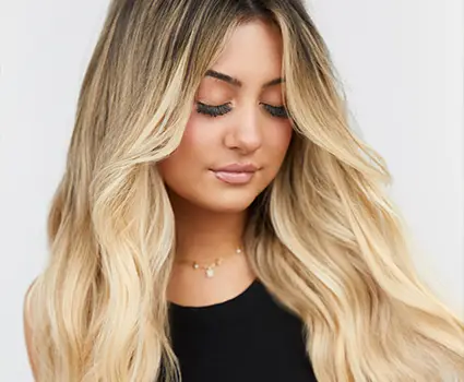 - Paris Parker Aveda Salons and Spas - Baton Rouge, New Orleans, Mandeville, LA A woman with long, wavy blonde balayage hair wearing a black top and a delicate gold necklace looks down with a neutral facial expression. - Paris Parker Aveda Salons and Spas - Baton Rouge, New Orleans, Mandeville, LA