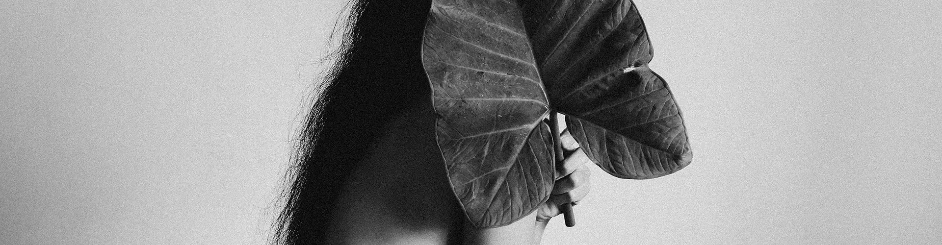 - Paris Parker Aveda Salons and Spas - Baton Rouge, New Orleans, Mandeville, LA A person with a fresh haircut stands with their back to the camera, holding a large leaf in front of their bare upper body. The black and white image exudes a sense of minimalist style. - Paris Parker Aveda Salons and Spas - Baton Rouge, New Orleans, Mandeville, LA