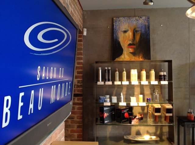 - Paris Parker Aveda Salons and Spas - Baton Rouge, New Orleans, Mandeville, LA Interior of a salon and day spa with a blue "Salon du Beau Monde" sign, shelves of hair products, and a painted portrait of a face hanging on a gray wall—perfect for balayage or a relaxing facial. - Paris Parker Aveda Salons and Spas - Baton Rouge, New Orleans, Mandeville, LA