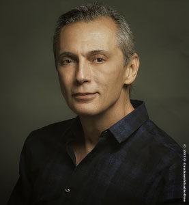 - Paris Parker Aveda Salons and Spas - Baton Rouge, New Orleans, Mandeville, LA A middle-aged man with short gray hair, styled in a balayage technique, wearing a dark collared shirt poses against a plain, dark background. - Paris Parker Aveda Salons and Spas - Baton Rouge, New Orleans, Mandeville, LA