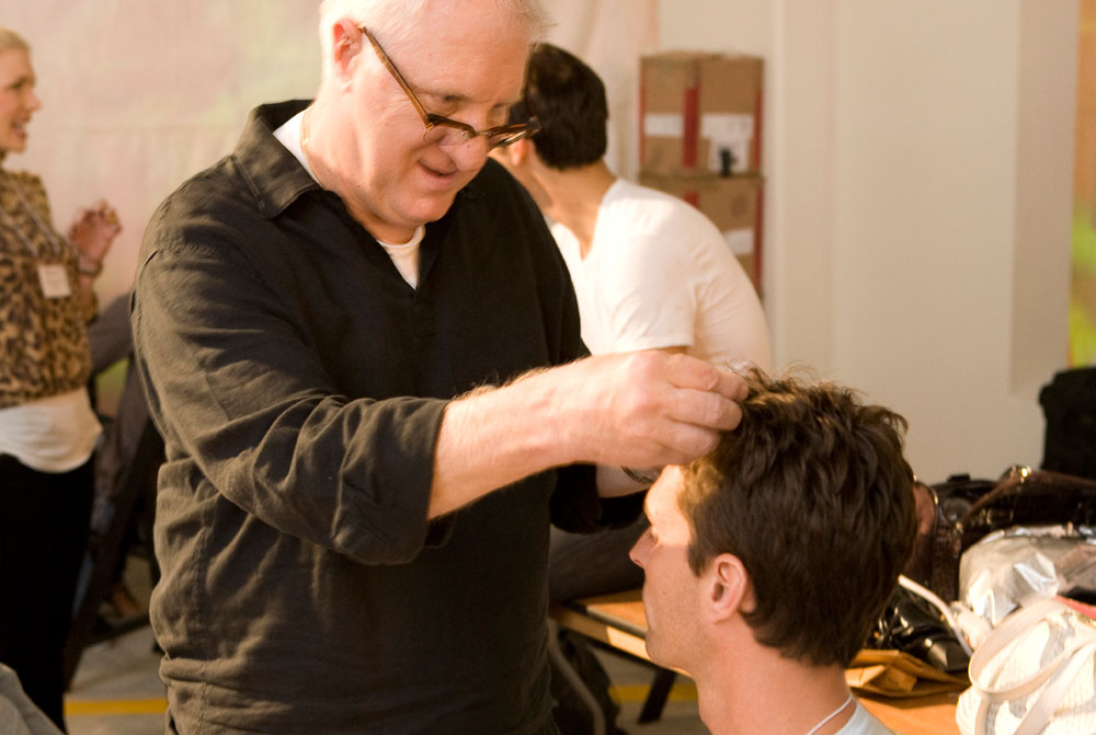 - Paris Parker Aveda Salons and Spas - Baton Rouge, New Orleans, Mandeville, LA A man standing styles the hair of a seated man in a well-lit indoor setting, suggesting a professional haircut. Other people and boxes are visible in the background, evoking the atmosphere of a busy day spa. - Paris Parker Aveda Salons and Spas - Baton Rouge, New Orleans, Mandeville, LA
