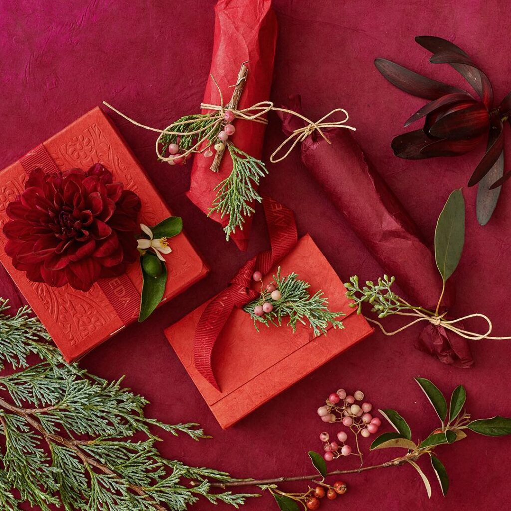 - Paris Parker Aveda Salons and Spas - Baton Rouge, New Orleans, Mandeville, LA Red and orange gifts wrapped with ribbon, twine, greenery, and berries are arranged on a red surface—perfect inspiration for thoughtful presents after a relaxing Aveda Salon facial or haircut. - Paris Parker Aveda Salons and Spas - Baton Rouge, New Orleans, Mandeville, LA