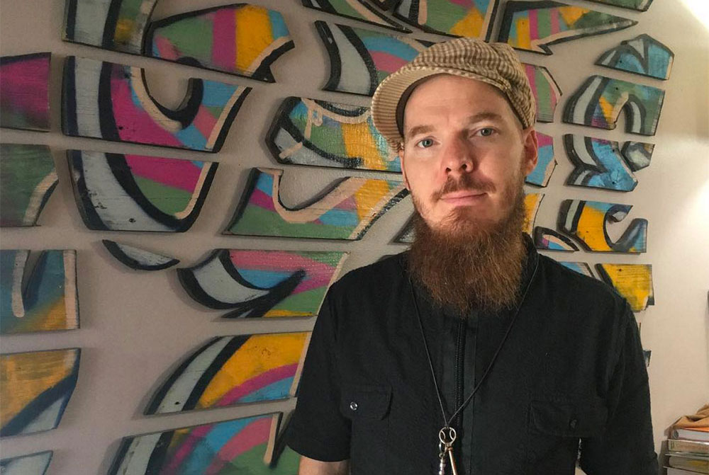 - Paris Parker Aveda Salons and Spas - Baton Rouge, New Orleans, Mandeville, LA Man with a long beard and cap stands in front of a wall with colorful abstract artwork made from separate wooden pieces, evoking the creativity often found in a modern hair salon or day spa. - Paris Parker Aveda Salons and Spas - Baton Rouge, New Orleans, Mandeville, LA