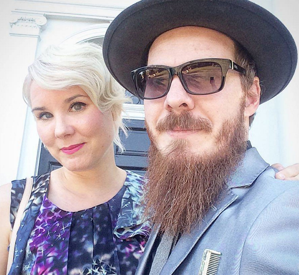 - Paris Parker Aveda Salons and Spas - Baton Rouge, New Orleans, Mandeville, LA A woman with a fresh haircut from an Aveda Salon stands next to a bearded man wearing sunglasses, a hat, and a gray suit; both are looking at the camera. - Paris Parker Aveda Salons and Spas - Baton Rouge, New Orleans, Mandeville, LA