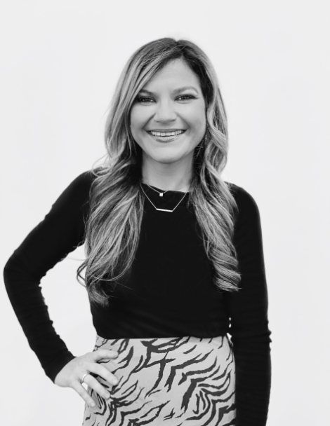 - Paris Parker Aveda Salons and Spas - Baton Rouge, New Orleans, Mandeville, LA A woman with a fresh Aveda Salon haircut, long wavy hair, wearing a black top and patterned skirt, stands smiling with one hand on her hip against a plain white background. - Paris Parker Aveda Salons and Spas - Baton Rouge, New Orleans, Mandeville, LA