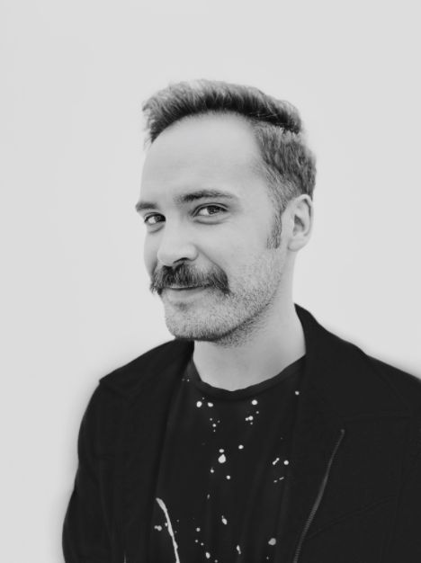 - Paris Parker Aveda Salons and Spas - Baton Rouge, New Orleans, Mandeville, LA Man with short hair and a mustache wearing a black jacket and patterned shirt, looking at the camera against a plain white background, showcasing a fresh look inspired by an Aveda Salon. - Paris Parker Aveda Salons and Spas - Baton Rouge, New Orleans, Mandeville, LA