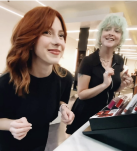 - Paris Parker Aveda Salons and Spas - Baton Rouge, New Orleans, Mandeville, LA Two women with balayage-dyed hair—one red, one mint green—smile while standing at a makeup counter in a well-lit store. - Paris Parker Aveda Salons and Spas - Baton Rouge, New Orleans, Mandeville, LA
