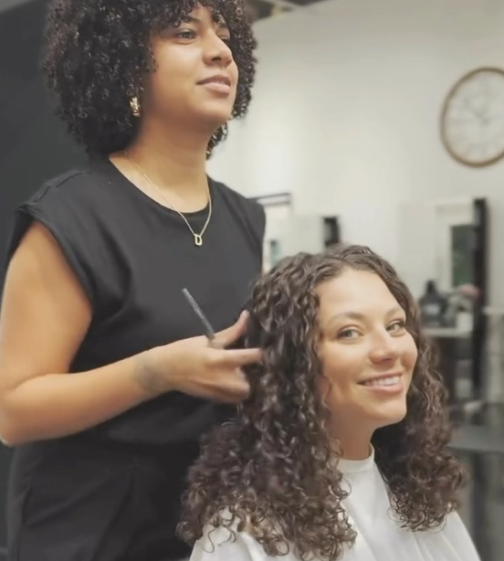 - Paris Parker Aveda Salons and Spas - Baton Rouge, New Orleans, Mandeville, LA A hairstylist stands behind a seated client with curly hair, preparing for a haircut, while the client smiles at the camera in a vibrant salon setting. - Paris Parker Aveda Salons and Spas - Baton Rouge, New Orleans, Mandeville, LA