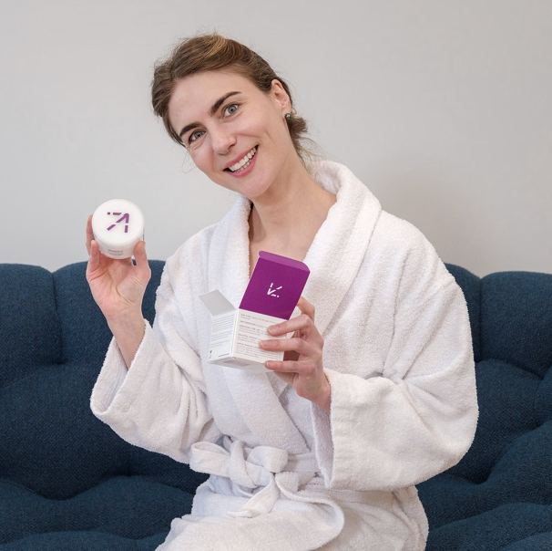- Paris Parker Aveda Salons and Spas - Baton Rouge, New Orleans, Mandeville, LA A woman in a white bathrobe sits on a blue couch, smiling and holding a round container and an open purple box with skincare products, as if she just returned from a hair salon after her fresh haircut or balayage treatment. - Paris Parker Aveda Salons and Spas - Baton Rouge, New Orleans, Mandeville, LA