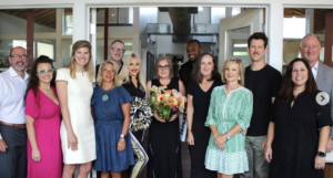 - Paris Parker Aveda Salons and Spas - Baton Rouge, New Orleans, Mandeville, LA A group of twelve adults poses indoors at an Aveda Salon, with one woman in the center holding a bouquet of flowers. Everyone is standing and smiling at the camera. - Paris Parker Aveda Salons and Spas - Baton Rouge, New Orleans, Mandeville, LA