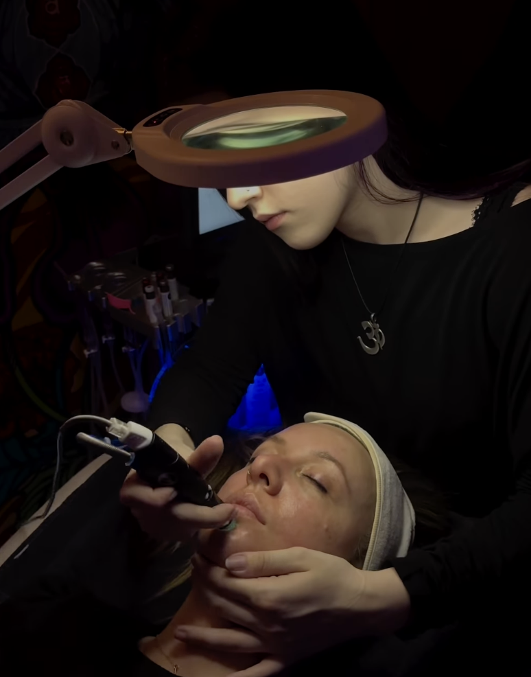- Paris Parker Aveda Salons and Spas - Baton Rouge, New Orleans, Mandeville, LA An aesthetician at an Aveda Salon uses a handheld device for a facial treatment on a client lying under a magnifying lamp, creating a relaxing day spa experience. - Paris Parker Aveda Salons and Spas - Baton Rouge, New Orleans, Mandeville, LA