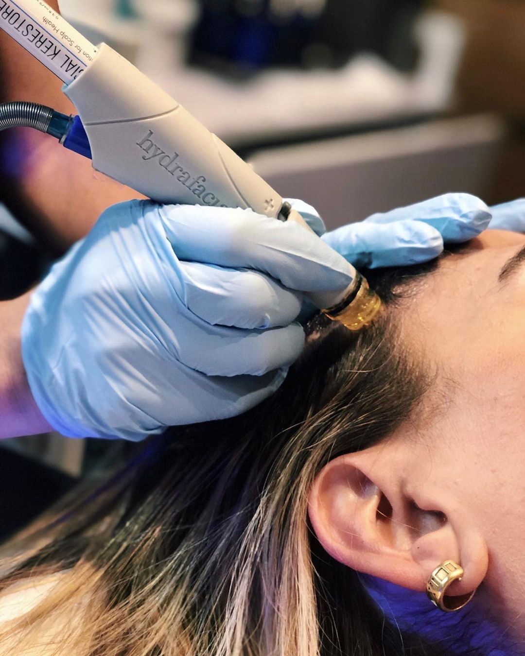 - Paris Parker Aveda Salons and Spas - Baton Rouge, New Orleans, Mandeville, LA A person receives a HydraFacial treatment on their forehead by a professional wearing blue gloves at an Aveda Salon and Day Spa. - Paris Parker Aveda Salons and Spas - Baton Rouge, New Orleans, Mandeville, LA
