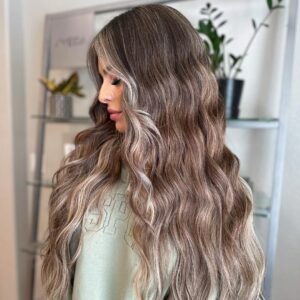 - Paris Parker Aveda Salons and Spas - Baton Rouge, New Orleans, Mandeville, LA Woman with long, wavy, light brown hair featuring blonde highlights stands indoors in front of potted plant shelves at a chic Aveda Salon and Day Spa, radiating natural beauty and relaxation. - Paris Parker Aveda Salons and Spas - Baton Rouge, New Orleans, Mandeville, LA