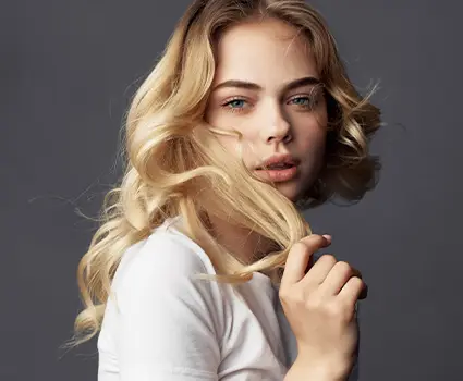 - Paris Parker Aveda Salons and Spas - Baton Rouge, New Orleans, Mandeville, LA Young woman with long, wavy blonde hair wearing a white shirt, holding a strand of her hair, and looking directly at the camera against a plain gray background, showcasing her fresh haircut from an Aveda Salon. - Paris Parker Aveda Salons and Spas - Baton Rouge, New Orleans, Mandeville, LA