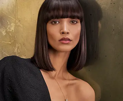 - Paris Parker Aveda Salons and Spas - Baton Rouge, New Orleans, Mandeville, LA A woman with straight, shoulder-length dark hair and blunt bangs poses against a textured gold wall, wearing a black off-shoulder top and a necklace, radiating the fresh glow of a facial at an Aveda Salon & Day Spa. - Paris Parker Aveda Salons and Spas - Baton Rouge, New Orleans, Mandeville, LA