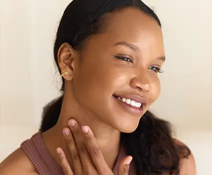 - Paris Parker Aveda Salons and Spas - Baton Rouge, New Orleans, Mandeville, LA A woman with smooth skin smiles softly while touching her neck, her hair styled to perfection as if fresh from an Aveda Salon, wearing a sleeveless top and small earrings against a plain background. - Paris Parker Aveda Salons and Spas - Baton Rouge, New Orleans, Mandeville, LA