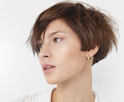 - Paris Parker Aveda Salons and Spas - Baton Rouge, New Orleans, Mandeville, LA Woman with short, tousled brown hair looking to the side, showcasing a fresh haircut, wearing a light-colored top and a small gold hoop earring. - Paris Parker Aveda Salons and Spas - Baton Rouge, New Orleans, Mandeville, LA