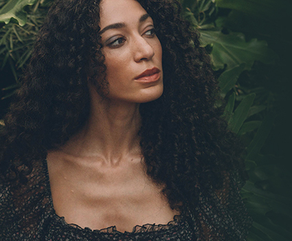 - Paris Parker Aveda Salons and Spas - Baton Rouge, New Orleans, Mandeville, LA Woman with long balayage curly hair wearing a dark patterned top looks to the side, standing in front of green leafy plants. - Paris Parker Aveda Salons and Spas - Baton Rouge, New Orleans, Mandeville, LA