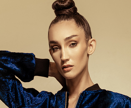 - Paris Parker Aveda Salons and Spas - Baton Rouge, New Orleans, Mandeville, LA Woman with a topknot hairstyle, fresh from an Aveda Salon haircut, wears a blue textured jacket and poses with one hand behind her neck against a plain beige background. - Paris Parker Aveda Salons and Spas - Baton Rouge, New Orleans, Mandeville, LA