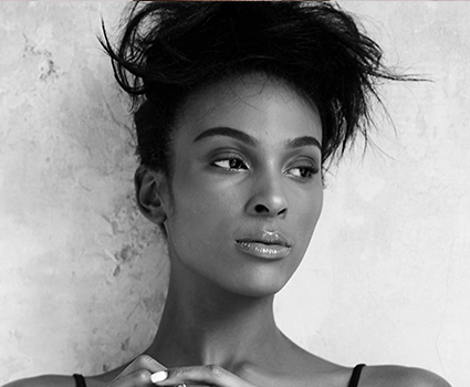 - Paris Parker Aveda Salons and Spas - Baton Rouge, New Orleans, Mandeville, LA Black and white photo of a woman with a fresh haircut, styled hair, and clear skin—perfect for a hair salon or facial promotion. She wears a tank top and stands against a textured wall, gazing slightly to her left. - Paris Parker Aveda Salons and Spas - Baton Rouge, New Orleans, Mandeville, LA