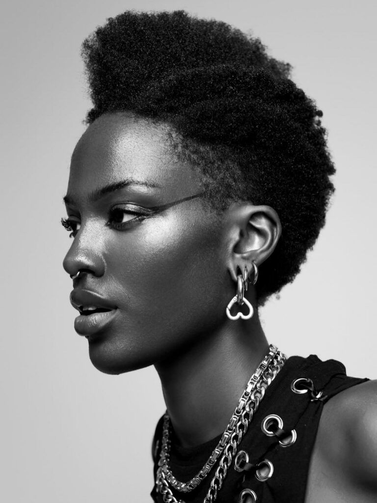 - Paris Parker Aveda Salons and Spas - Baton Rouge, New Orleans, Mandeville, LA Black and white profile portrait of a person with short, natural hair, wearing hoop earrings, a nose ring, and layered chain necklaces—capturing a chic look perfect for a modern hair salon. - Paris Parker Aveda Salons and Spas - Baton Rouge, New Orleans, Mandeville, LA