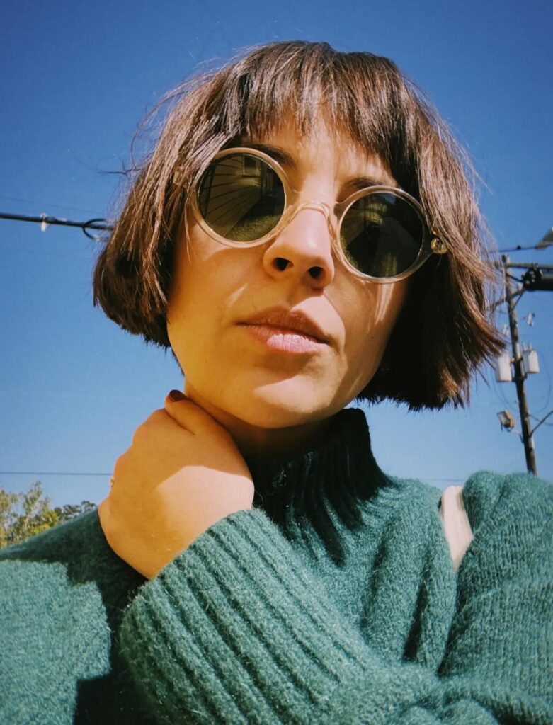 - Paris Parker Aveda Salons and Spas - Baton Rouge, New Orleans, Mandeville, LA A person with short brown hair wearing round sunglasses and a green sweater stands outside under a clear blue sky, looking refreshed as if just back from a facial at an Aveda Salon. - Paris Parker Aveda Salons and Spas - Baton Rouge, New Orleans, Mandeville, LA