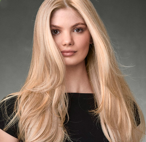 Aveda Hair Color - Paris Parker Aveda Salons and Spas - Baton Rouge, New Orleans, Mandeville, LA Woman with long, straight, blonde hair wearing a black top, facing forward against a plain gray background. - Paris Parker Aveda Salons and Spas - Baton Rouge, New Orleans, Mandeville, LA